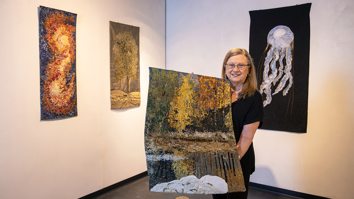 Dawn Murtaugh showing off her quilted textile titled Steam Boat Springs while standing in front of 3 other quilted textiles depicting the cosmos, a tree, and a jelly fish