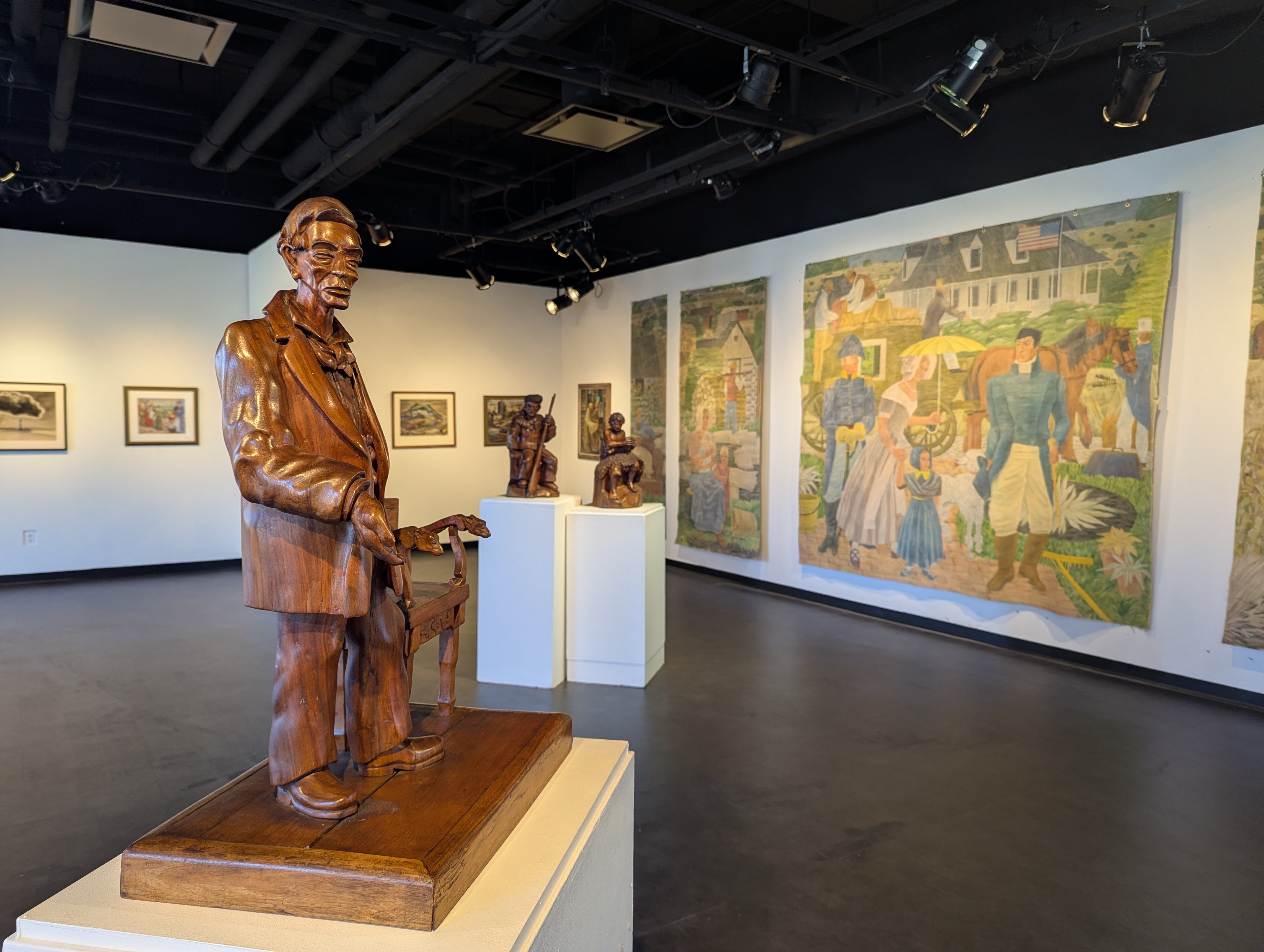 view of for the people exhibit showing fred myers moris library lincoln wood carving infront of framed art work and hanging murals all from new deal era